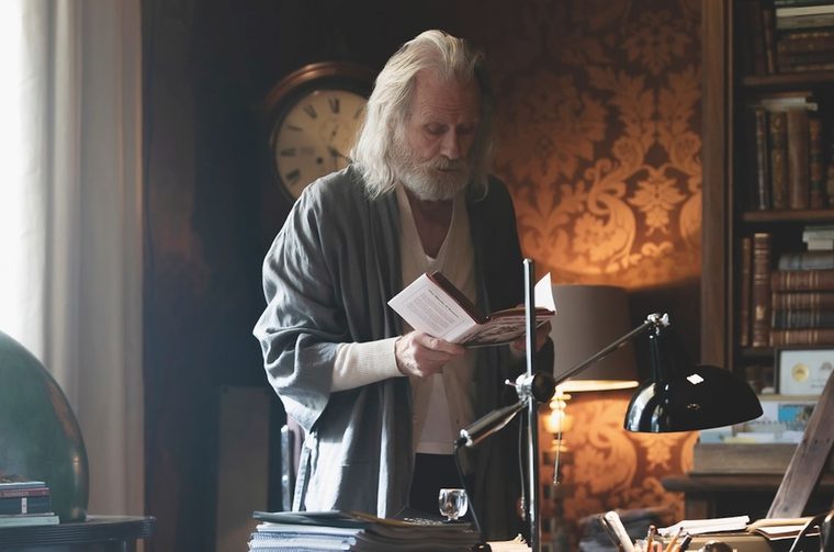 An elderly man with long white hair and a full white beard stands in a book-lined study, reading a small open book he holds in both hands. He wears a white shirt under a gray cardigan and long gray robe. The warm, dimly lit room features a bookshelf packed with leather-bound volumes, an antique wall clock, ornate gold-patterned wallpaper, a black articulated desk lamp, a globe, and stacks of papers and books on a cluttered desk in the foreground.
