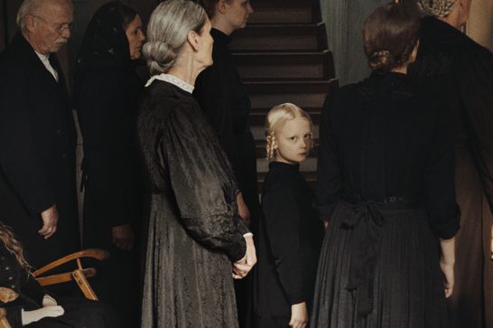 We see a collection of men and women of different ages wearing black early 20th century clothing. They all face away from the camera apart from a small blonde haired child who stands amongst them, looking directly at the camera with a sombre expression. We can see a staircase behind them.