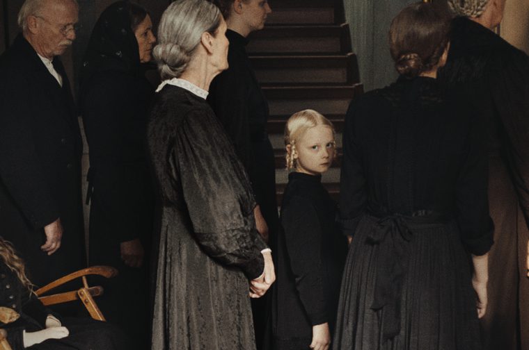 We see a collection of men and women of different ages wearing black early 20th century clothing. They all face away from the camera apart from a small blonde haired child who stands amongst them, looking directly at the camera with a sombre expression. We can see a staircase behind them.