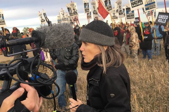A female journalist in her fifties holding a microphone reports in front of a large crowd of protesters holding signs reading 'Defend the Sacred,' 'Water Is Life,' 'Greed Kills,' and similar messages, at an outdoor demonstration on grassland. A camera operator with a boom microphone films in the foreground.