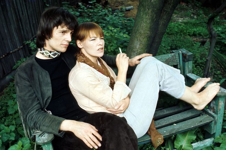 A young man with messy black hair and wearing dark clothes and a choker necklace with a diamond pattern sits on a bench in a wooded area. Next to him, leaning on his left arm, sits an older middle aged woman with chestnut coloured hair holding a cigarette. She has her bare feet up on the bench and is wearing blue jeans, a pale blouse and a brown scarf.