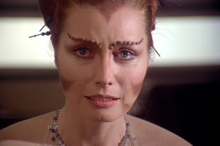 A close-up of a middle aged woman with an elaborate updo held by ornamental hairpins, with textured ridged eyebrows and subtle forehead prosthetics suggesting an alien species. She has tearful eyes and a distressed expression, wearing a beaded, multi-strand necklace and a pale blush-toned top, set against a softly lit interior background.