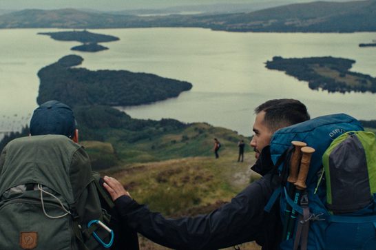 Two hikers with backpacks stand on a hilltop overlooking a large lake dotted with wooded islands. One man, facing away in a green pack and navy cap, rests a hand on the shoulder of his companion, who wears a teal backpack with trekking poles and looks out at the view. Rolling green hills and misty mountains stretch into the distance.