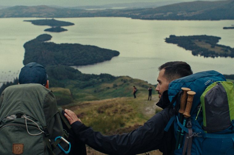 Two hikers with backpacks stand on a hilltop overlooking a large lake dotted with wooded islands. One man, facing away in a green pack and navy cap, rests a hand on the shoulder of his companion, who wears a teal backpack with trekking poles and looks out at the view. Rolling green hills and misty mountains stretch into the distance.