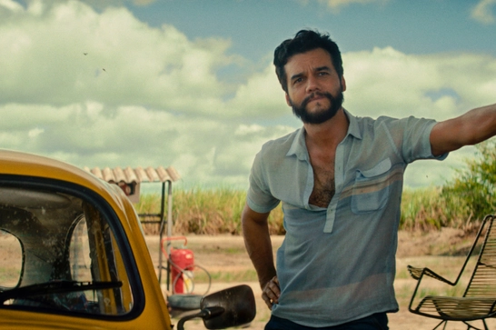 We see a Brazilian man in his 40s with a beard wearing a light blue short-sleeved shirt, leaning on a wall and standing outdoors in what looks like a rural or agricultural setting. There's a yellow vintage car beside him, and sugarcane fields are visible in the background under a partly cloudy sky.