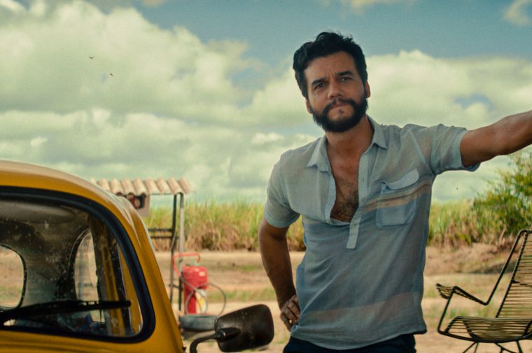 We see a Brazilian man in his 40s with a beard wearing a light blue short-sleeved shirt, leaning on a wall and standing outdoors in what looks like a rural or agricultural setting. There's a yellow vintage car beside him, and sugarcane fields are visible in the background under a partly cloudy sky.