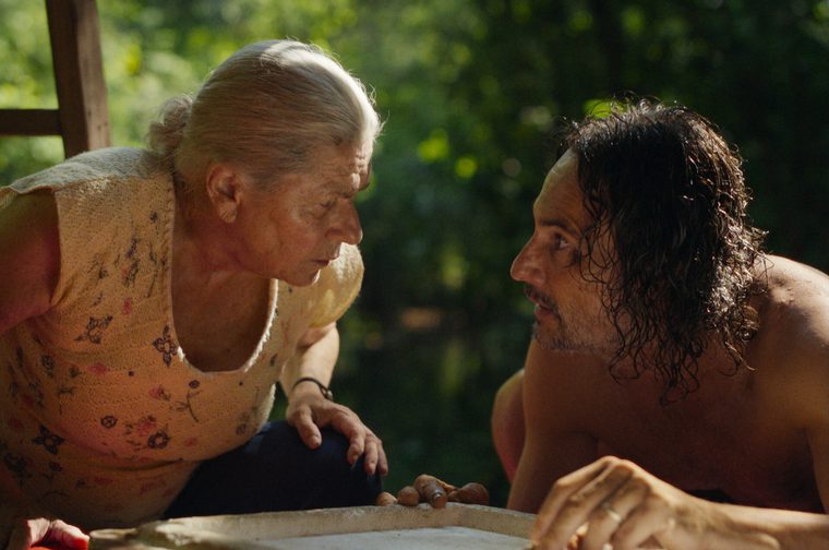 An elderly woman with white hair pulled back, wearing a floral sleeveless top, leans forward intently across a table toward a shirtless middle-aged man with long, wet, dark curly hair and a short beard. The two face each other in close proximity, suggesting a tense or intimate exchange. They are outdoors or in an open-air setting, with lush green trees visible in the soft-focus background. Warm, dappled natural light fills the scene.