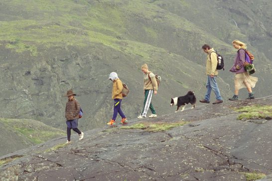 We see a family - a father, mother and three children - moving along a high rocky outcrop, a black and white dog by their feet. They each have rucksacks on and we can see patches of green on the rocks as they move down the mountain.