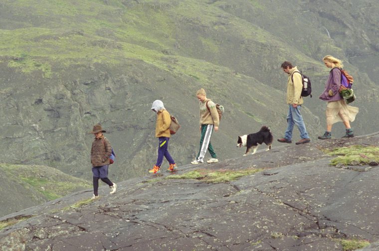 We see a family - a father, mother and three children - moving along a high rocky outcrop, a black and white dog by their feet. They each have rucksacks on and we can see patches of green on the rocks as they move down the mountain.