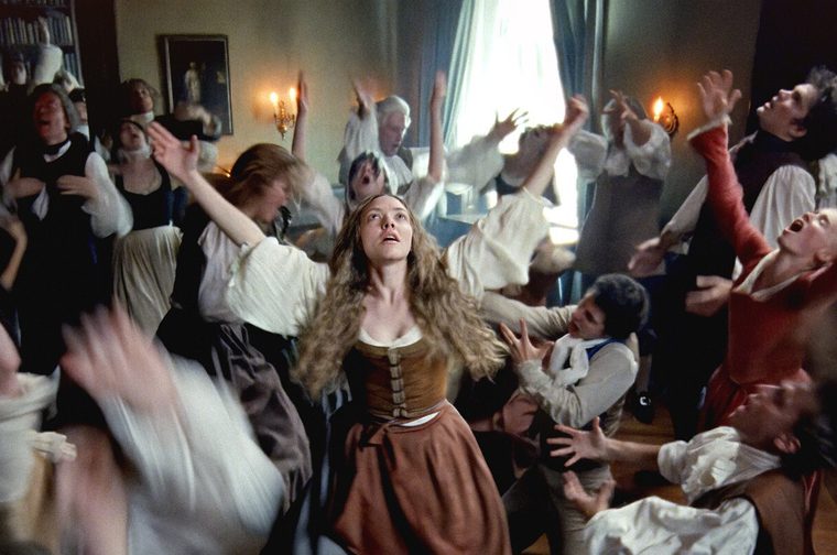 A woman in her forties dressed in 18th century clothing holds her hands out to the ceiling of the room she is standing in, her eyes lifted up to the heavens in ecastasy. Around her lots of men and women dance and flail around her, some of their images blurry. We see paintings and candelabras on the walls.