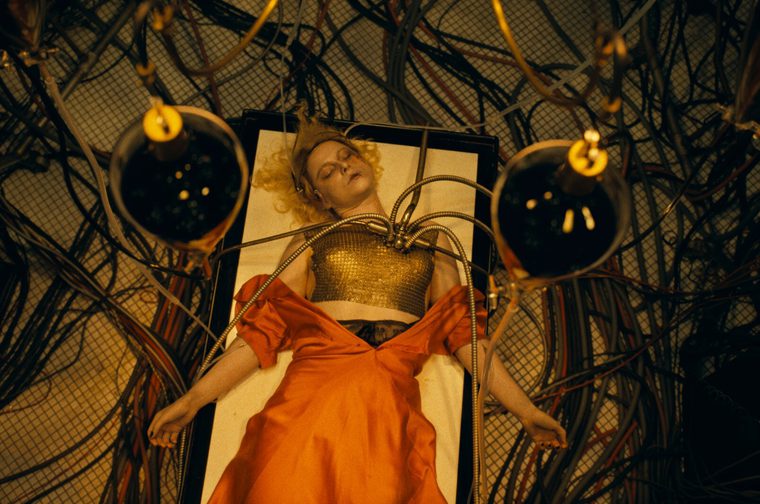 A woman with blonde hair (Jessie Buckley) lies unconscious on a flat surface, wearing a gold metallic top and a voluminous orange skirt. Metallic tubes and cables are attached to her torso, spreading outward like mechanical tendrils. Two large spherical devices flank her on either side. The background is filled with a tangle of wires and cables in a dimly lit, industrial setting. The image has a dramatic gothic aesthetic.
