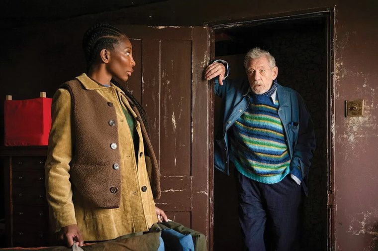 A dimly lit interior scene showing two people facing each other in what appears to be a worn, dark-walled room. On the left, a Black woman with long braided hair wears a yellow corduroy jacket over a brown fleece vest, holding a bundle of fabric at her side. On the right, an elderly white man with white hair and a beard leans against a dark wooden door frame, dressed in a colorful blue-and-green striped sweater beneath an open blue jacket and navy trousers. A red object hangs on the wall in the background. The atmosphere is tense and theatrical.