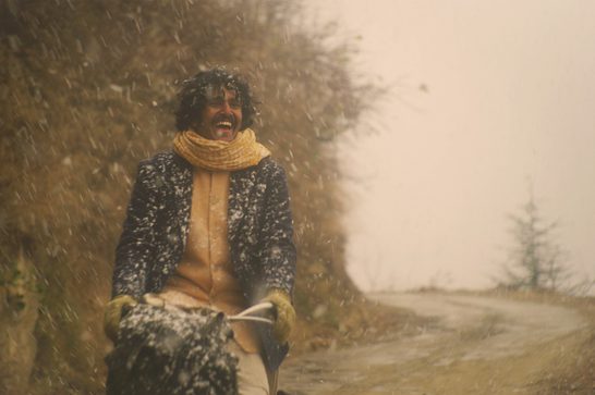 A young Indian man with curly dark hair and a mustache laughs joyfully while riding a horse-drawn sled or cart through falling snow on a dirt road. He wears a dark speckled wool coat dusted with snow, a mustard-yellow scarf and sweater, and olive gloves, gripping the reins. Bare trees and dry autumn brush line the road on the left, while the right side opens to a hazy, pale sky with a few sparse evergreen trees in the distance.