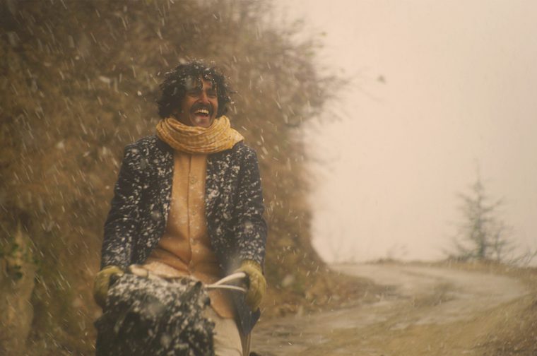 A young Indian man with curly dark hair and a mustache laughs joyfully while riding a horse-drawn sled or cart through falling snow on a dirt road. He wears a dark speckled wool coat dusted with snow, a mustard-yellow scarf and sweater, and olive gloves, gripping the reins. Bare trees and dry autumn brush line the road on the left, while the right side opens to a hazy, pale sky with a few sparse evergreen trees in the distance.