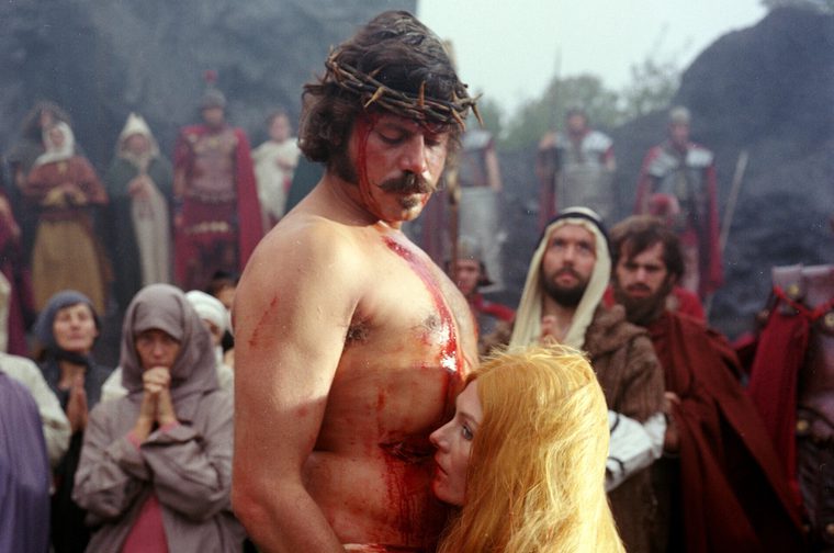 A bloodied man wearing a crown of thorns stands bare-chested, his torso streaked with fake blood, while a blonde-haired woman leans her head against his chest. Behind them, a crowd of extras in period robes, headscarves, and Roman-style armor and capes looks on, some with hands clasped in prayer.