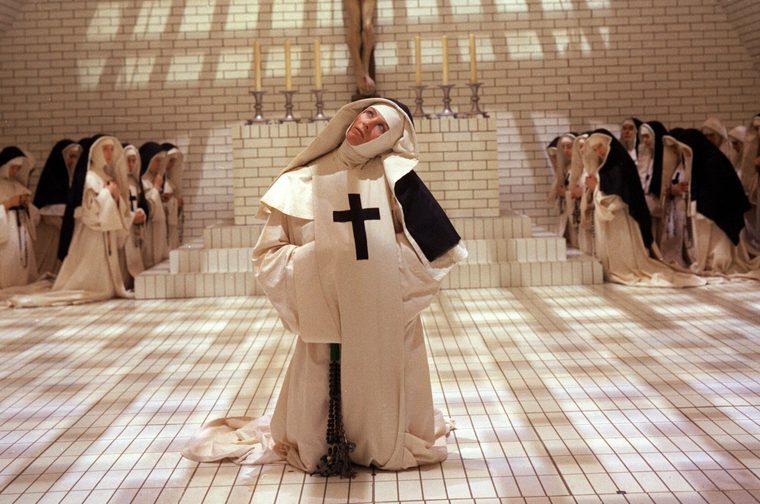 A nun in a cream habit with a black cross on her scapular kneels at the center of a stark white-tiled chapel, gazing upward with a rapt expression, a long rosary hanging in front of her. Rows of kneeling nuns in matching habits flank a tiled altar topped with a crucifix and six candles, while window shadows form a grid across the floor and walls.