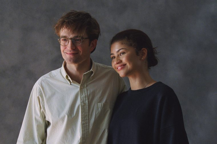 We see two people posing together in front of a grey studio backdrop. On the left is a man with light brown hair and round glasses, wearing a light cream/white button-up shirt, smiling warmly. On the right is a woman with dark hair pulled back, wearing a dark navy/black top, also smiling. They appear relaxed and comfortable together.