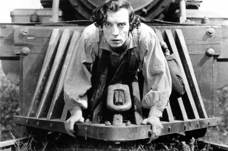 A black-and-white still from a silent-era film. A young man (Buster Keaton) with dark, curly hair and an intense, wide-eyed expression crouches low on the front cowcatcher of a large steam locomotive, gripping the metal frame with both hands. He wears a loose striped shirt and appears to be in a moment of high tension or peril, staring directly into the camera. The massive iron body of the train looms behind him.