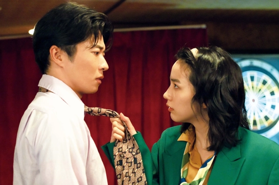 A Japanese woman in a green blazer grabs a Japanese man's patterned tie, both facing each other with intense expressions. There's a red curtain and a dartboard visible in the background.