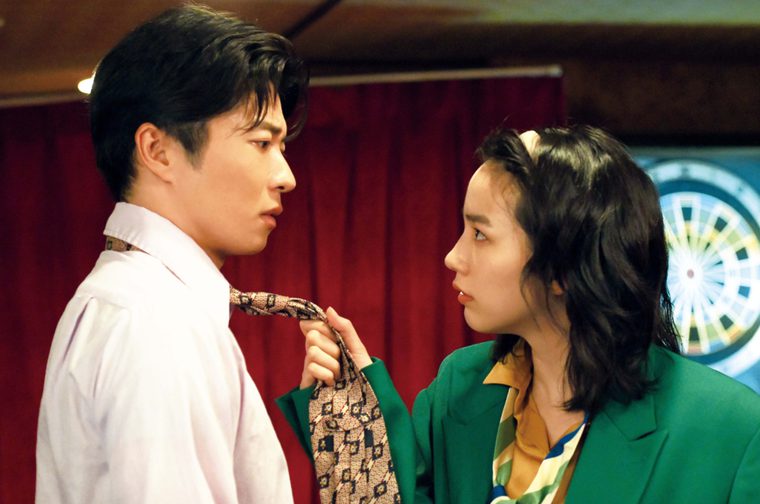 A Japanese woman in a green blazer grabs a Japanese man's patterned tie, both facing each other with intense expressions. There's a red curtain and a dartboard visible in the background.
