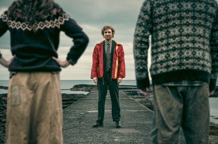 A red-haired man in a red-and-yellow rain jacket, dress shirt, and tie stands on a stone jetty facing the camera with a tense, uncertain expression, flanked in soft focus by two figures in traditional fisherman's sweaters with their backs turned toward him. The overcast sea and sky stretch out behind him.