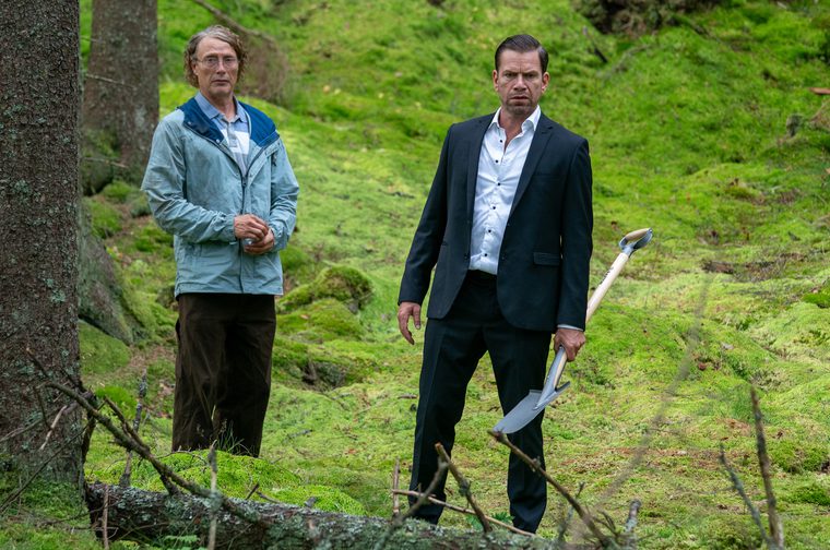 Two men stand in a lush, moss-covered forest. On the left, a slender man wearing glasses and a light blue jacket looks ahead with a neutral expression. On the right, a broad-shouldered man in a black suit and open-collar white shirt holds a shovel at his side, looking directly at the camera with an intense expression.