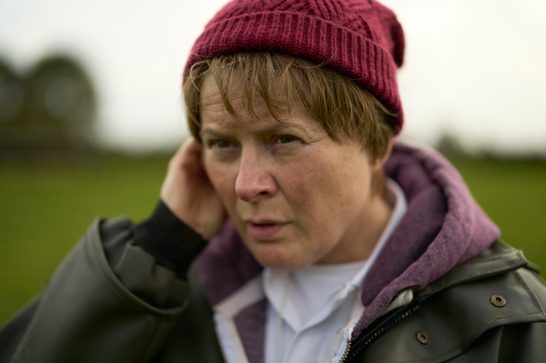 Close-up of a middle aged woman with short, light-brown hair and a burgundy knitted beanie, one hand raised to her ear. She looks at the camera with a furrowed, serious expression, wearing a dark olive waxed jacket over a purple hoodie and white collared shirt, against a blurred green field and overcast sky.