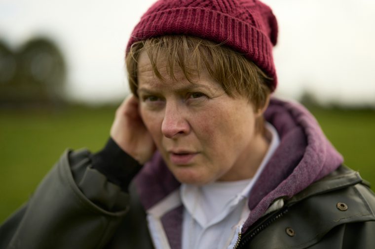 Close-up of a middle aged woman with short, light-brown hair and a burgundy knitted beanie, one hand raised to her ear. She looks at the camera with a furrowed, serious expression, wearing a dark olive waxed jacket over a purple hoodie and white collared shirt, against a blurred green field and overcast sky.