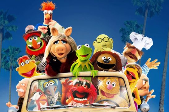The image shows a large group of beloved Muppet characters piled onto and around a classic car, set against a sunny blue sky with palm trees — very much in the style of The Muppet Movie promotional artwork. Recognizable characters include Miss Piggy and Kermit the Frog front and center, along with Fozzie Bear (driving), Animal, Gonzo, Beaker, Scooter, Rowlf, Swedish Chef, Dr. Teeth, Statler & Waldorf, and many others