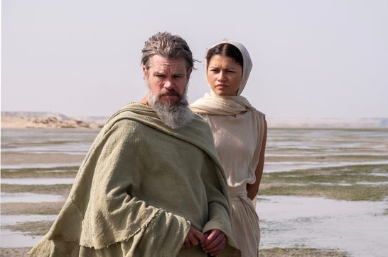Two figures stand on a vast, arid coastal flat under a pale, hazy sky. In the foreground, an older man with silver-streaked hair and a full grey beard wears a coarse, draped olive-green robe, his expression stern and weathered. Behind him, a younger woman in cream-coloured linen — a sleeveless dress and a loose head wrap — looks forward with a furrowed brow. The landscape stretches behind them: shallow tidal mudflats reflecting muted light, with low rocky ridges visible in the far distance. The overall palette is desaturated and sun-bleached.