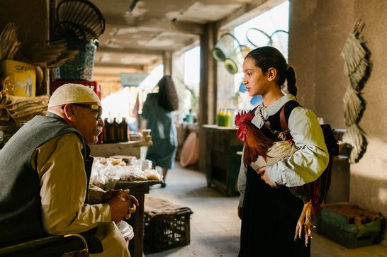A young Iraqi girl in a school uniform — white blouse and dark pinafore — carries a large rooster in her arms while facing an older seated man wearing a kufi cap and glasses. They appear to be in conversation inside a covered market or bazaar, with goods and food items visible on tables behind the man.