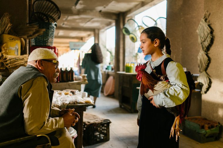 A young Iraqi girl in a school uniform — white blouse and dark pinafore — carries a large rooster in her arms while facing an older seated man wearing a kufi cap and glasses. They appear to be in conversation inside a covered market or bazaar, with goods and food items visible on tables behind the man.
