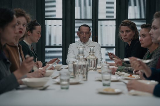 We see a man in a white military-style uniform seated at the head of a long white-draped table, staring directly at the camera with a stern expression. Several women are seated on both sides of the table, eating from bowls and plates with uneasy, somber expressions. The room has tall windows with slatted blinds in the background, casting a cold, institutional atmosphere.