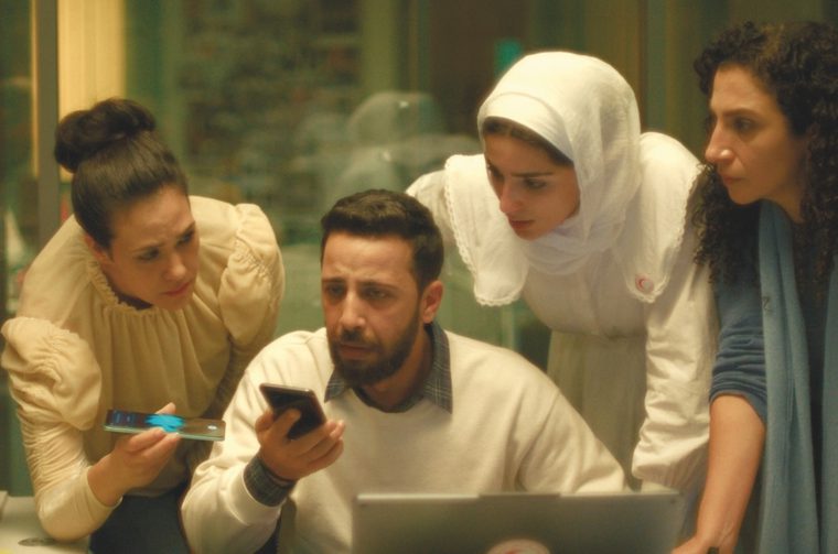 The image shows four people — three women and one man — huddled together in what appears to be an office or control room setting, all intently focused on a phone the man is holding. The man (center) is seated, wearing a white sweatshirt over a plaid shirt, holding a smartphone and looking at it with a serious, tense expression. The woman on the left is leaning in, wearing a cream/beige ruffled blouse with her dark hair up, also holding what looks like a phone or device, and peering at his screen with concern. The woman in the center-right is wearing a white hijab and a white uniform with a red crescent emblem — indicating she's a Red Crescent worker — leaning forward over a laptop. The woman on the far right has curly dark hair and is wearing a blue top, also leaning in and looking at the screen with a worried expression.