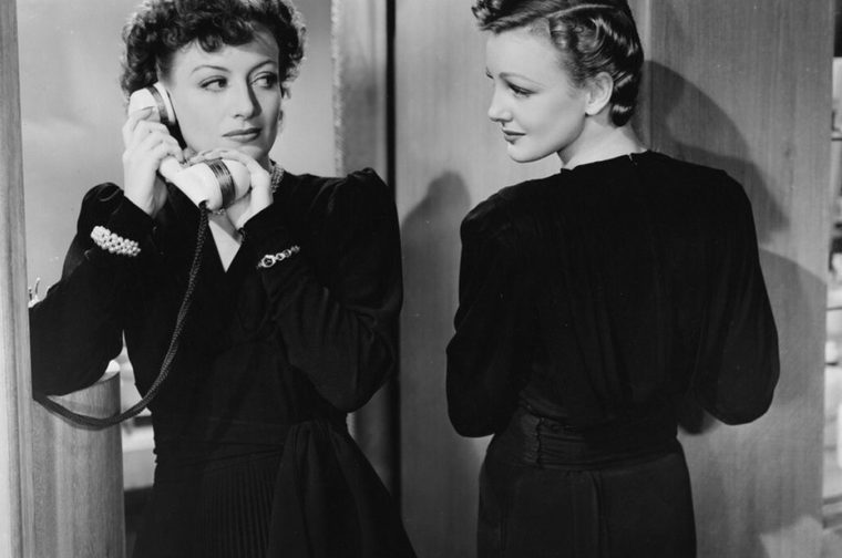 Black-and-white still from a 1930s-style film showing two white women in a wood-paneled room. On the left, a woman with short, curly dark hair holds a white telephone receiver to her ear with both hands, its cord trailing down. She wears a dark dress with puffed shoulders, a jeweled brooch, a pearl bracelet, and a bangle, and she looks sideways toward the other woman with a knowing, wary expression. On the right, a woman with sleek, sculpted waves pinned up on her head stands in profile, her back partly to the camera, wearing a dark dress with a wide, gathered waistband. She gazes back at the first woman with a composed, faintly cool look.