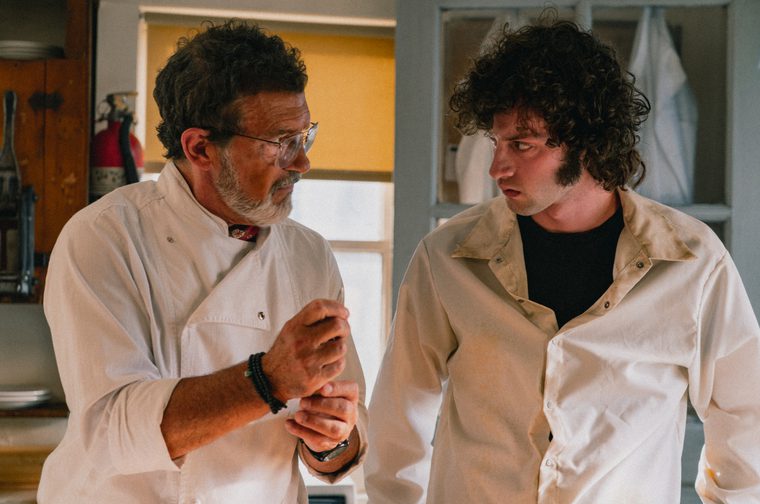 Two chefs in white coats stand close together in a kitchen, the older man on the left with curly gray-brown hair, glasses, and a beard focused on something in his hands, while the younger man with dark curly hair looks over at him intently.