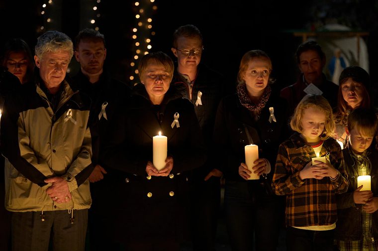 A group of adults and children stand together outdoors at night holding lit candles during a vigil. Several wear white ribbons pinned to their coats. Their expressions are solemn, and strings of small lights glow softly in the dark background.