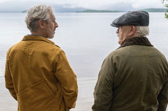 Two older men stand at the shore of a calm loch, their backs to the camera, facing each other in quiet conversation. The man on the left wears a mustard-yellow jacket; the man on the right wears an olive-green jacket and a grey flat cap. Misty hills and green treelines are visible across the water under an overcast sky.