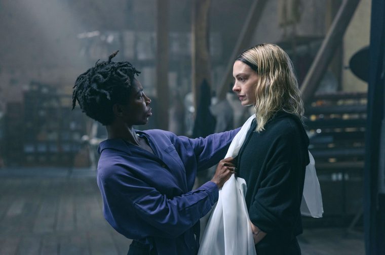 Two women face each other in a dimly lit, industrial-style interior space. One person, wearing a purple/blue shirt with short dreadlocks, reaches out and touches the chest of the other, who has shoulder-length blonde hair and wears a dark blazer with a white scarf. The mood is tense and intimate. Wooden beams and a staircase are visible in the blurred background.