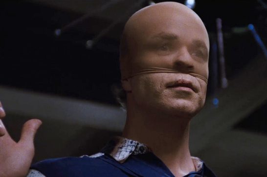 A bald male in his forties wearing a face-obscuring mask made of stretched, stitched material with stitching along the jaw and cheek, gestures upward with one hand while illuminated from a stage-like setting with dark rigging visible overhead.