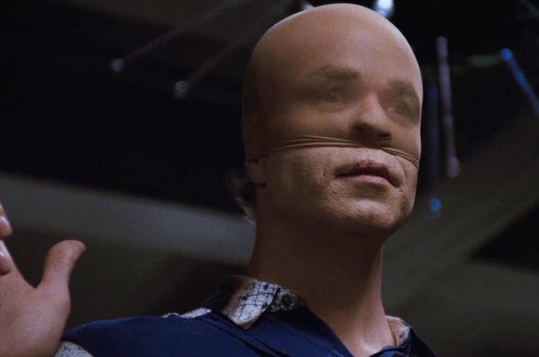 A bald male in his forties wearing a face-obscuring mask made of stretched, stitched material with stitching along the jaw and cheek, gestures upward with one hand while illuminated from a stage-like setting with dark rigging visible overhead.