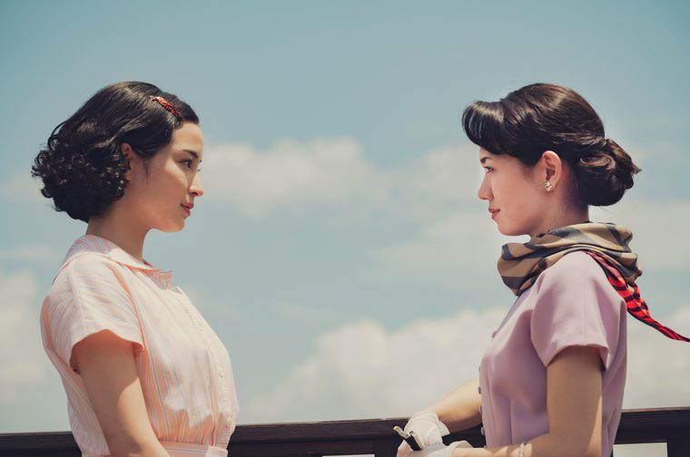 Two younger Japanese women in 1950s-style clothing face each other in profile against a bright blue sky with scattered clouds. The woman on the left wears a pale pink striped short-sleeve blouse and has dark curly hair with a decorative clip. The woman on the right has dark hair in an elegant updo and wears a soft pink short-sleeve dress with a patterned scarf tied loosely around her neck. Both women wear white gloves and rest their hands on a dark wooden railing