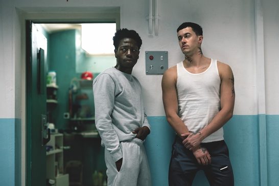 A young Black man stands next to a young white man in a prison corridor. The Black man is wearing a grey tracksuit and the white man is wearing a white vest with dark blue trousers. He also has tattoos across his arms and neck. They both wear sombre expressions. Behind them is an open door to what looks like a cleaning cupboard.