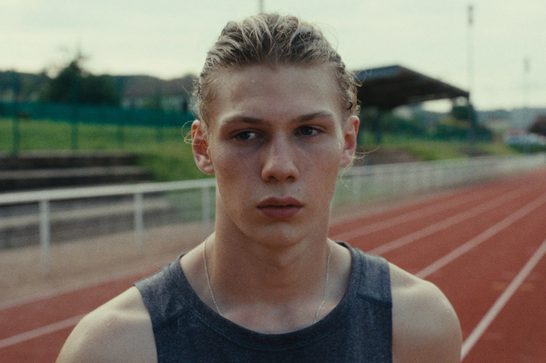 A young male athlete with damp, slicked-back blonde hair stands on a red running track, wearing a dark grey sleeveless top and a thin chain necklace. He gazes intensely into the distance with a serious expression. A sports stadium grandstand and green fencing are visible in the blurred background.