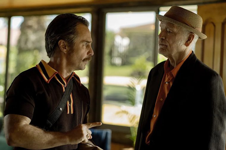 Two older white men talk in a sunlit room with large windows and honey-toned wood paneling. On the left, a man with slicked-back dark hair and a mustache wears a brown knit polo with orange-and-cream striped trim and a dark strap across his chest. He points with one finger as he speaks. On the right, an older man with white stubble and a straw fedora wears a dark blazer over an orange shirt and listens, looking at him intently. Blurred greenery and a lawn are visible through the windows behind them.