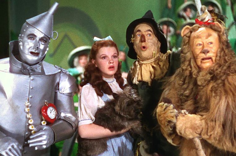 We see Dorothy (holding Toto) with her three companions in the Emerald City: the Tin Man (left), the Scarecrow (behind her), and the Cowardly Lion (right). This scene is from their audience with the Wizard, given with green-tinted lighting and awestruck expressions on their faces.