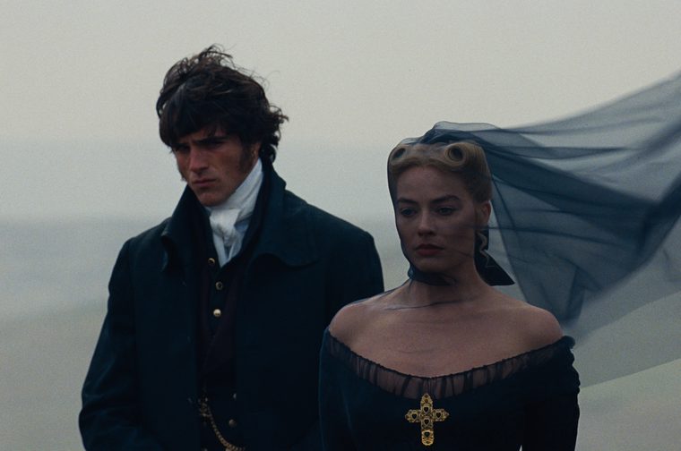 A man (Jacob Elordi) and woman (Margot Robbie) in period costume stand on a windswept Yorkshire moor. They both wear black, with the blonde haired woman wearing a black veil that billows in the wind behind her. She also has a gold cross on the front of her dress. They both wear sombre expression.