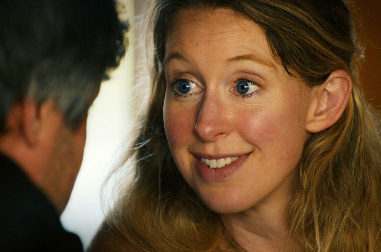 Close-up of a middle-aged white woman with long, tousled light brown hair, wide blue eyes, and an eager, slightly awkward smile. She gazes toward a person in the left foreground, seen from behind with dark, graying curly hair, out of focus. Warm, golden lighting fills the background.