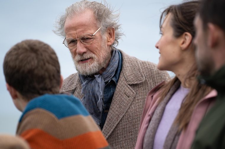 An elderly man with white hair, a white beard, and round glasses wearing a tweed blazer and blue scarf engages in conversation with a small group of people outdoors. A young person in a striped jumper stands to his left, and a woman in a pink top is visible to his right. The background is an overcast sky, suggesting a coastal or open-air setting.