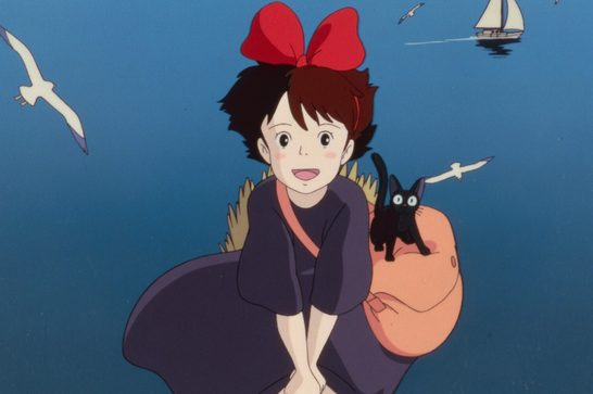 We see an animated shot of a young witch with short dark hair and a large red bow, flying on her broomstick over the ocean. She wears a purple dress and has a peach/salmon-colored bundle strapped to her broom, on which her black cat companion Jiji is perched, looking startled with wide eyes. Seagulls fly nearby and a sailboat is visible on the water in the background.